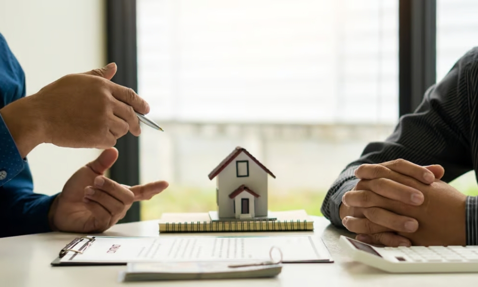 Two professionals sitting at table in front of window with paperwork and a small house