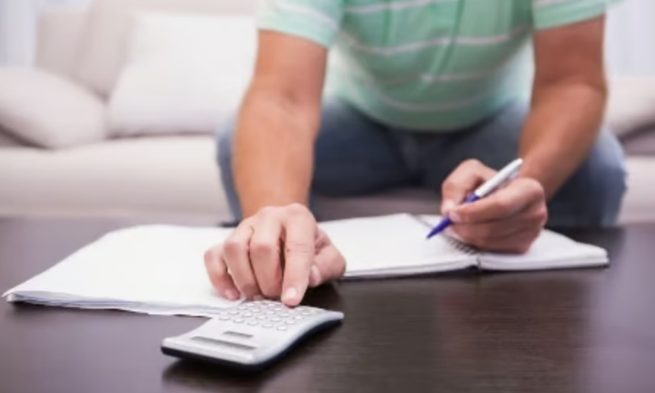 Man writing on white paper and using calculator on dark table, sitting on light colored couch