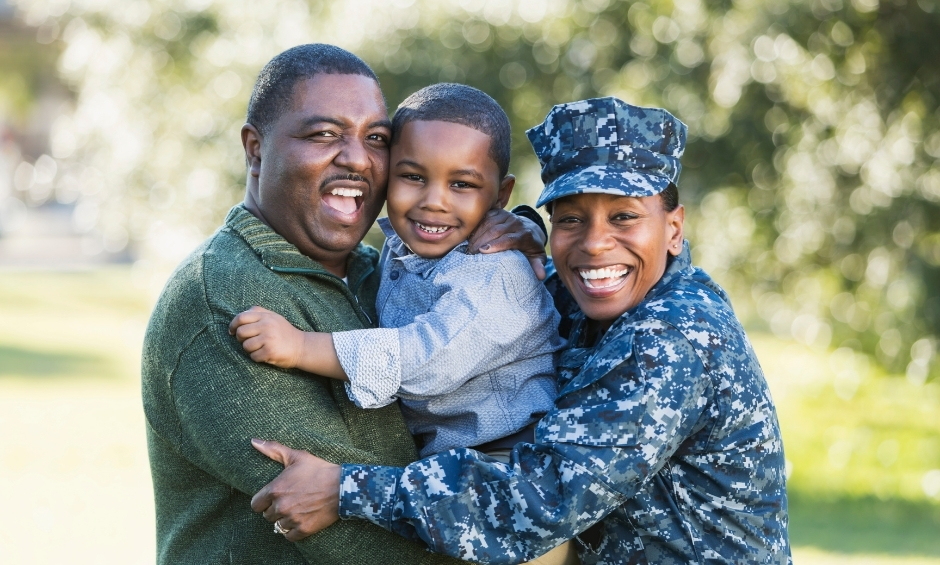 Couple holding a child, hugging. Male in green shirt, young boy in light shirt, woman in military uniform standing outside in front of trees