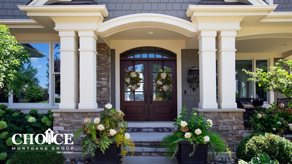 Gray stone home with cream pillars, arched entryway, stairs leading to dark doors, green trees and yellow flowers on green bushes