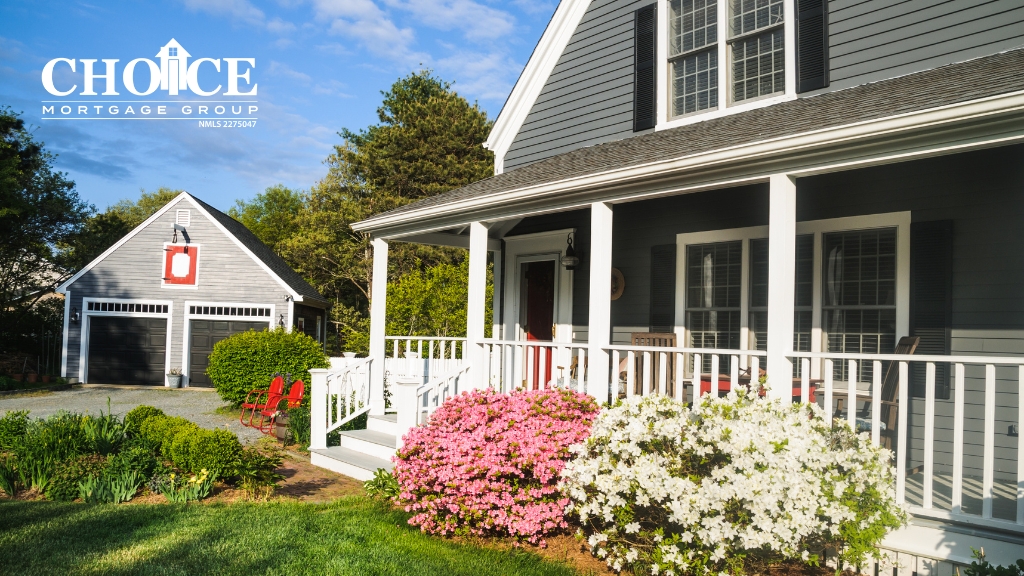 Gray craftsman style home with white fencing around porch, green grass, large white and pink flower bushes. blue skies