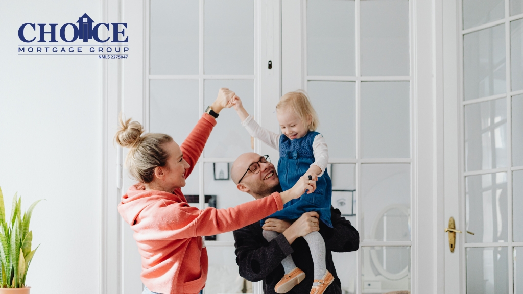 Woman with light hair in a bun wearing an orange sweatshirt holds hands with a child in a blue dress, while a bald man in a dark shirt holds the child, standing together in front of glass doors.