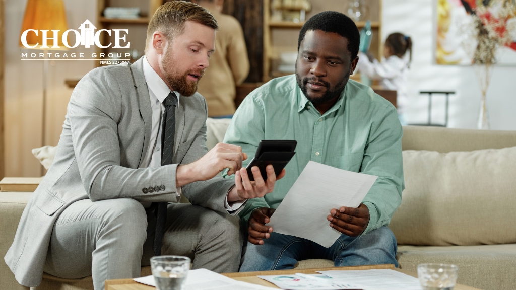 Two men sitting on beige couch with home setting in background. Man in gray suit with blue tie holding a calculator showing it to a man in green shirt holding a document.